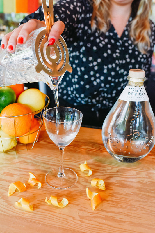 Classic Martini cocktail being poured into glass with citrus twists. Bottle of Freeland Spirits Dry Gin next to cocktail on bar at Freeland's tasting room & distillery in Portland, OR.