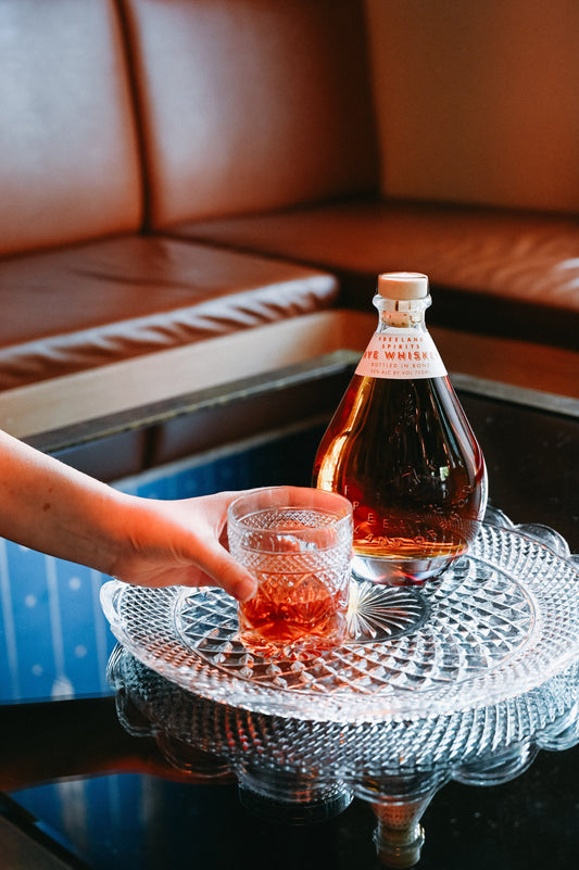 Freeland Spirits Rye Whiskey bottle on crystal serving platter next to sazerac cocktail. Hand reaching for cocktail from out of frame.