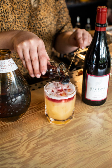 Mixologist preparing wine float for PDX State of Mind (New York Sour) cocktail, next to bottles of Freeland Bourbon and Elk Cove Pinot Noir