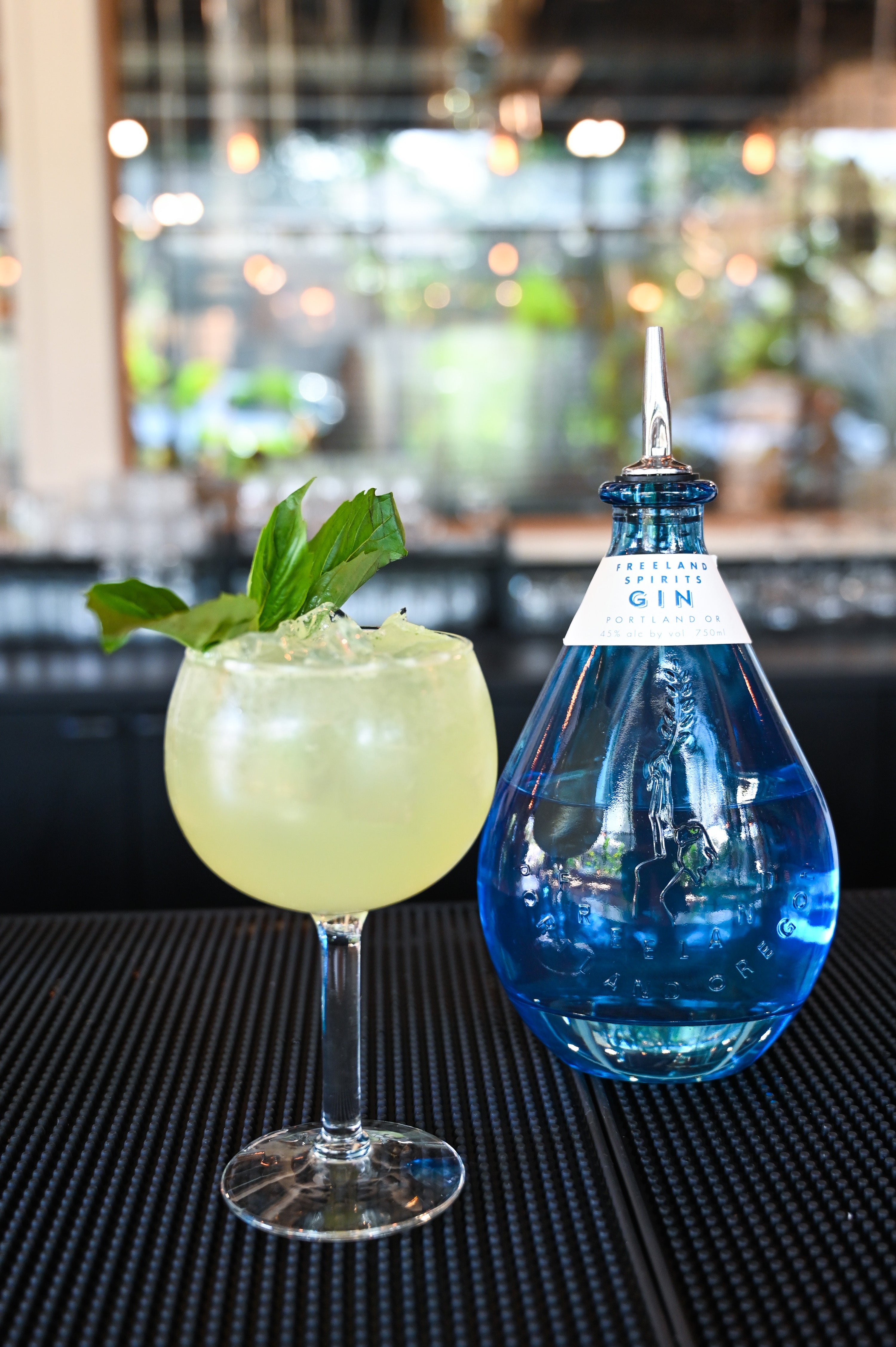 Bird's Eye View Gin & Tonic garnished with thai basil, on bar next to bottle of Freeland Gin