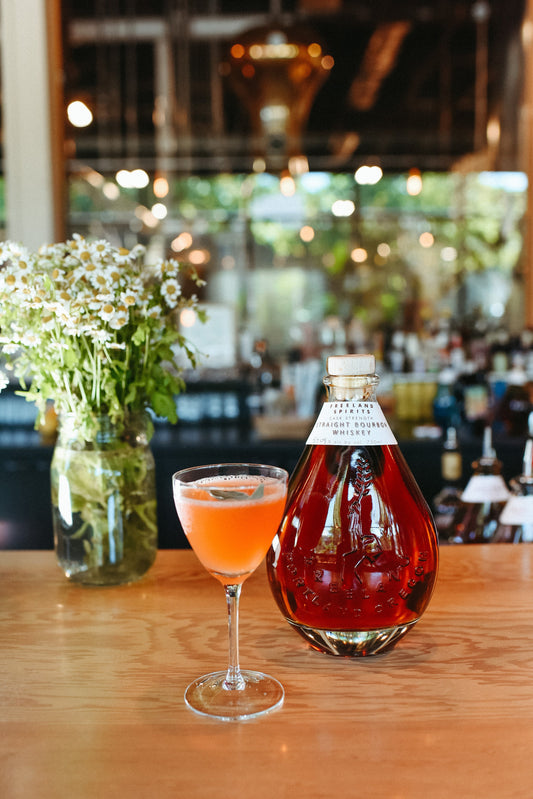 Freeland Spirits tasting room in Portland, OR. Paper Airplane cocktail (orange with sage garnish), next to vase of chamomille flowers and bottle of Freeland Cask Strength Whiskey Bourbon