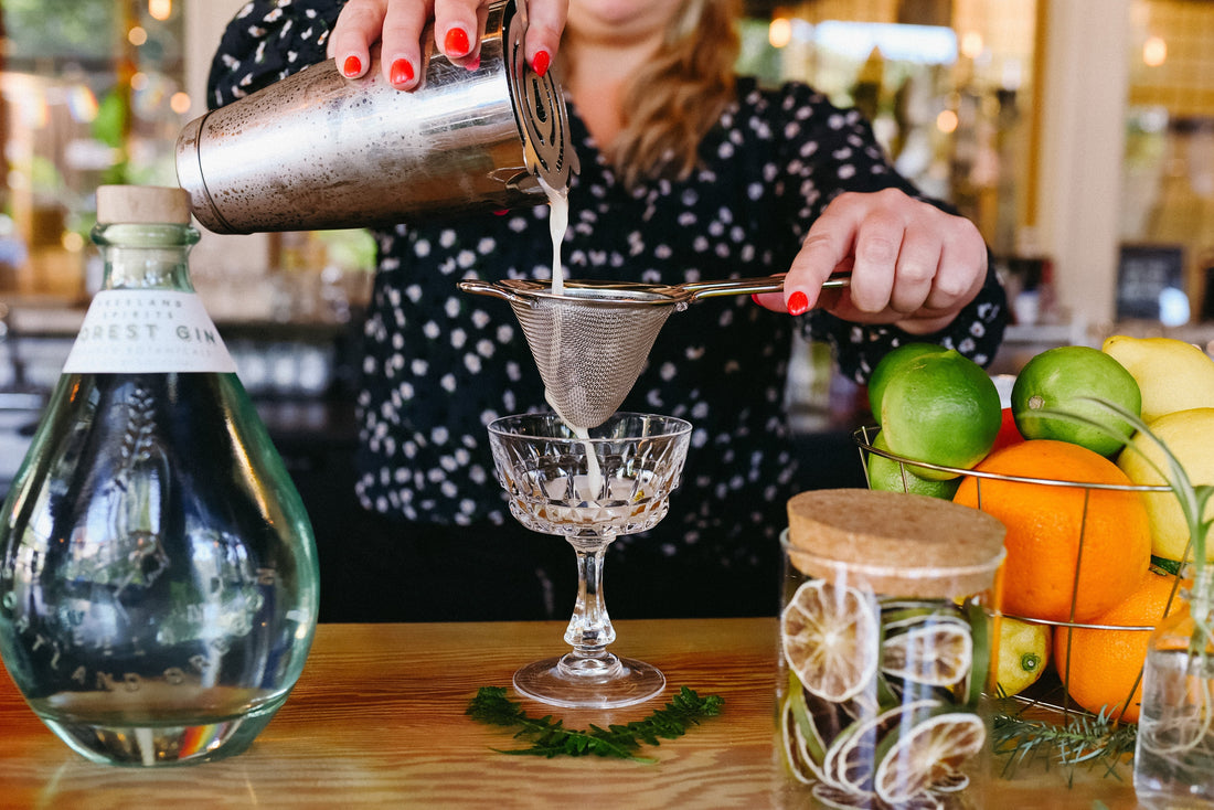 Into the Wild Freeland Spirits cocktail being prepared by a mixologist, Basket of citrus, jar of dried orange slices, and bottle of Freeland Forest Gin in a green bottle. 