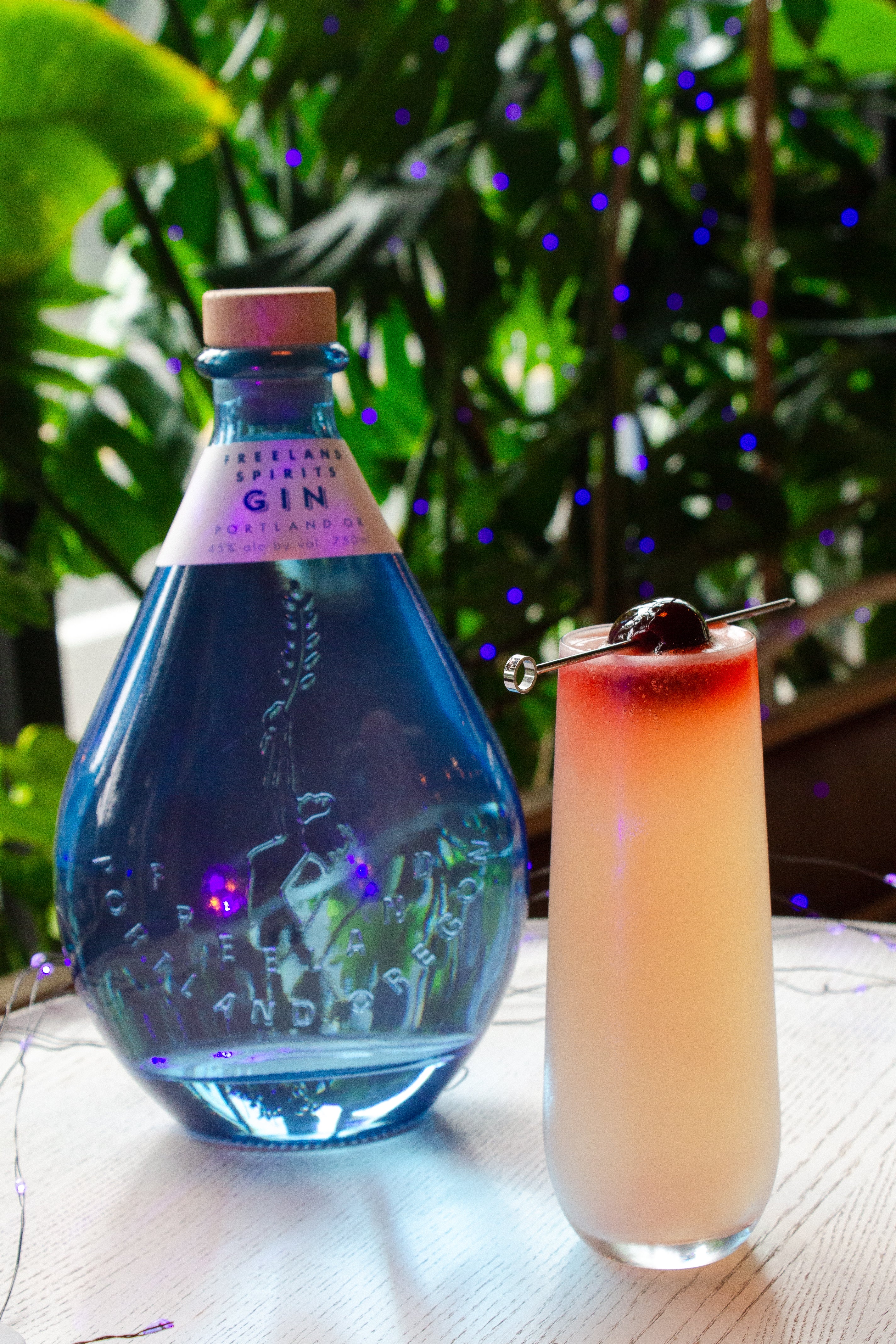 My Bloody Valentine bubbly cocktail garnished with cherry, next to bottle of Freeland Gin. Plants in background. 