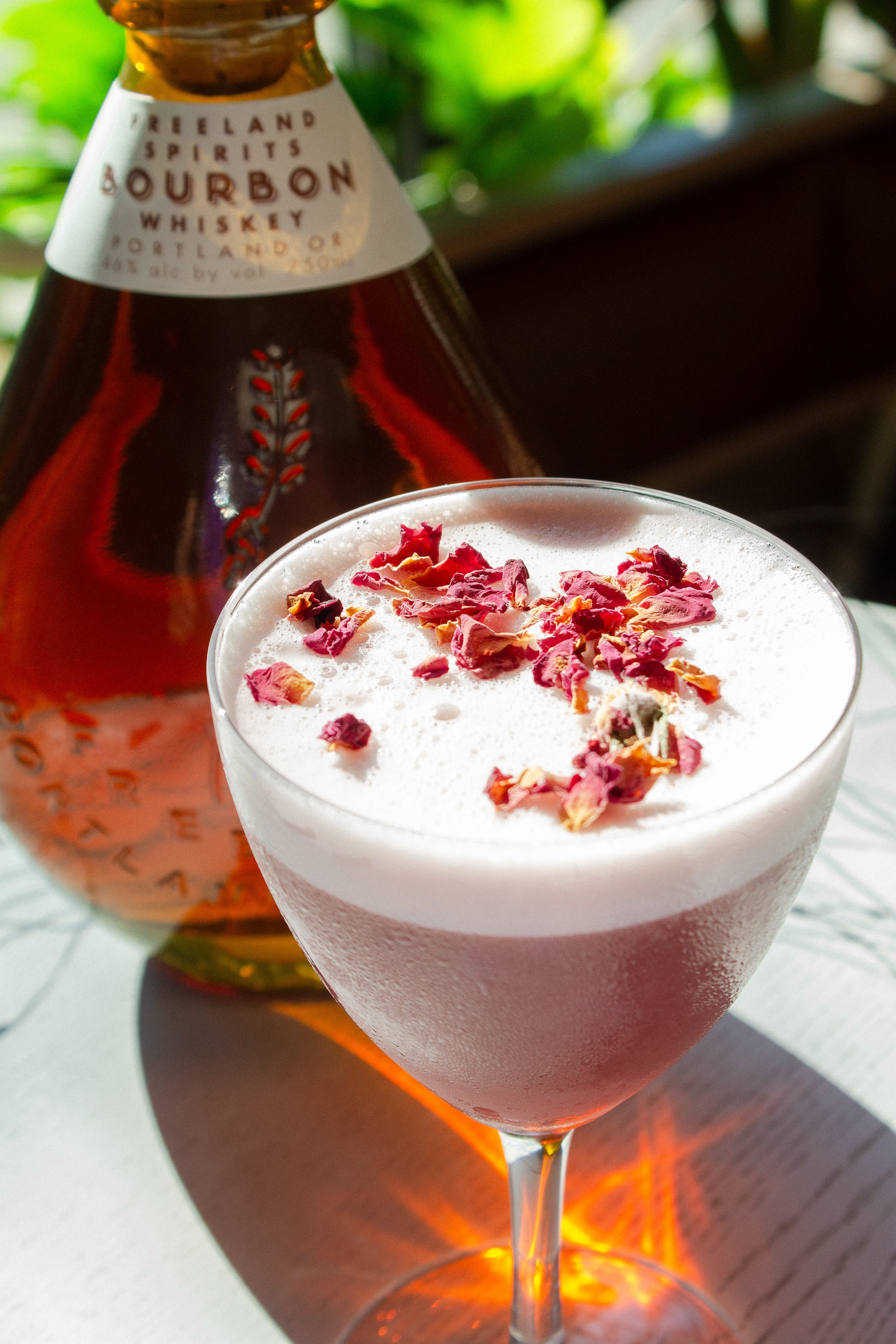 Mixologist adding dried rose petals to garnish Morticia & Gomez frothy Halloween cocktail, next to a bottle of Freeland Bourbon. 