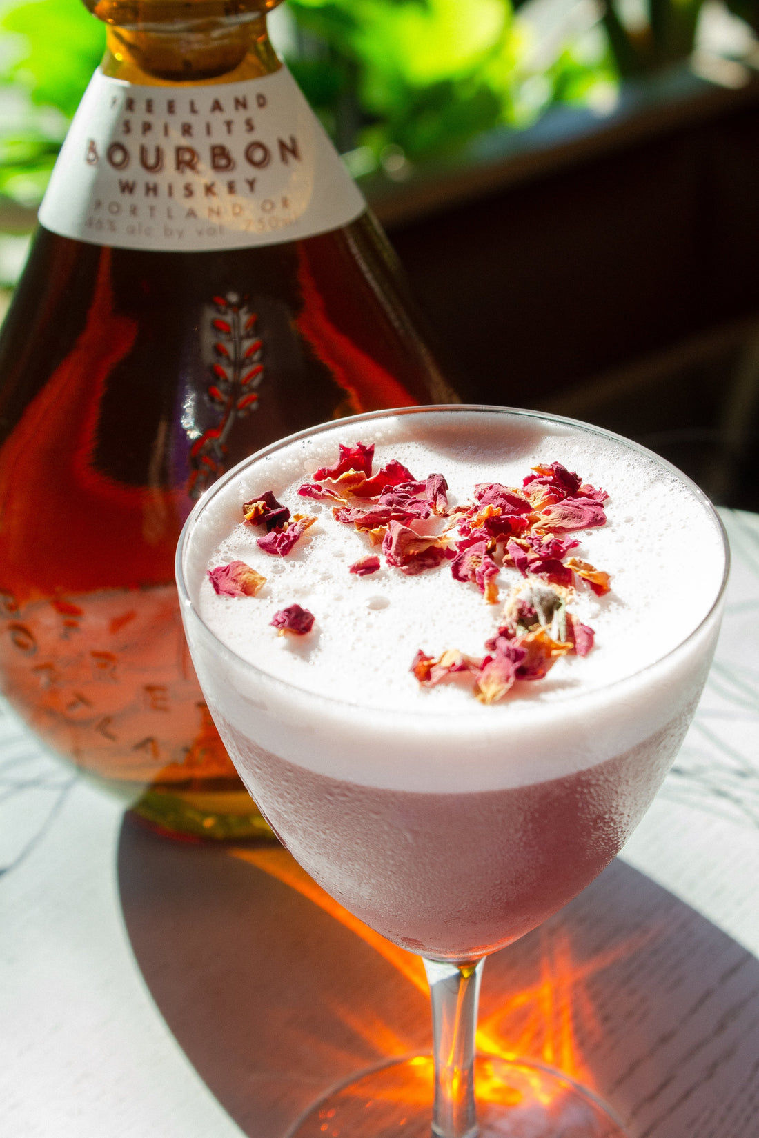 Mixologist adding dried rose petals to garnish Morticia & Gomez frothy Halloween cocktail, next to a bottle of Freeland Bourbon. 