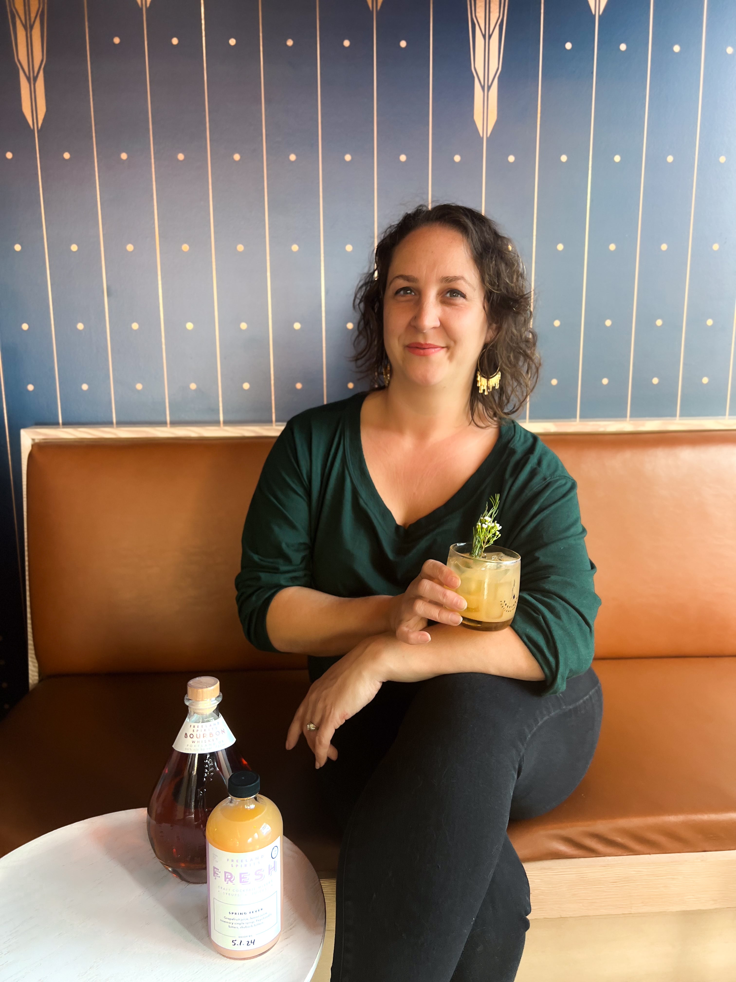 Heather Keisler Fornes, Executive Director of the Portland Fruit Tree Project at Freeland Spirits Tasting Room in Portland, OR holding a Spring Fever cocktail, next to a bottle of Freeland Bourbon