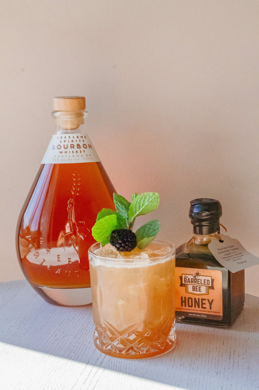 Cocktail special at Freeland Spirits for Black History Month. The Mrs. Margaret cocktail garnished with a blackberry and mint, next to bottle of Freeland Bourbon and the Barreled Bee honey. 