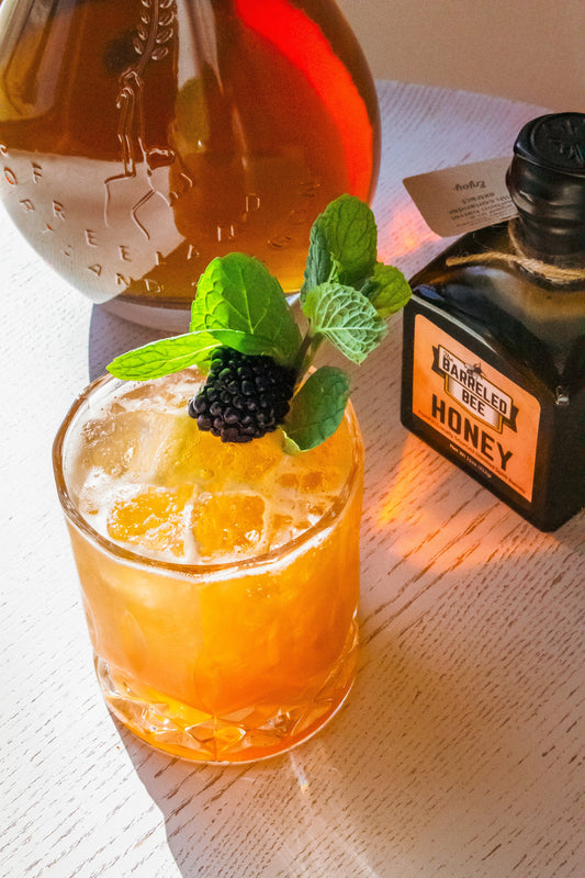 The Mrs. Margaret cocktail garnished with mint and blackberry, next to bottle of Barreled Bee honey and bottle of Freeland Spirits Bourbon in Portland, Oregon 