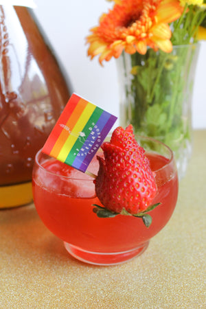 Strawberry Blvd - Pride cocktail at Freeland Spirits, garnished with a strawberry and Pride flag. Set next to a bottle of Bourbon and flowers.