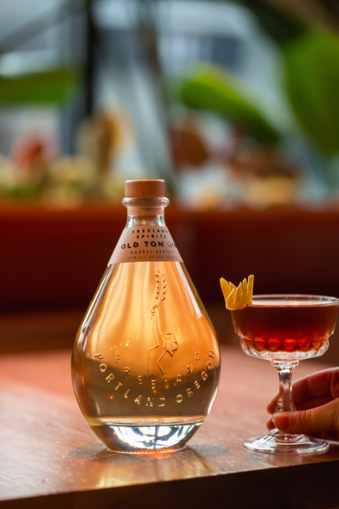 Bottle of Freeland Spirits Old Tom Gin next to Nic & Nora glass of a Martinez gin cocktail garnished with an orange peel, with a hand placing the cocktail on the table. 