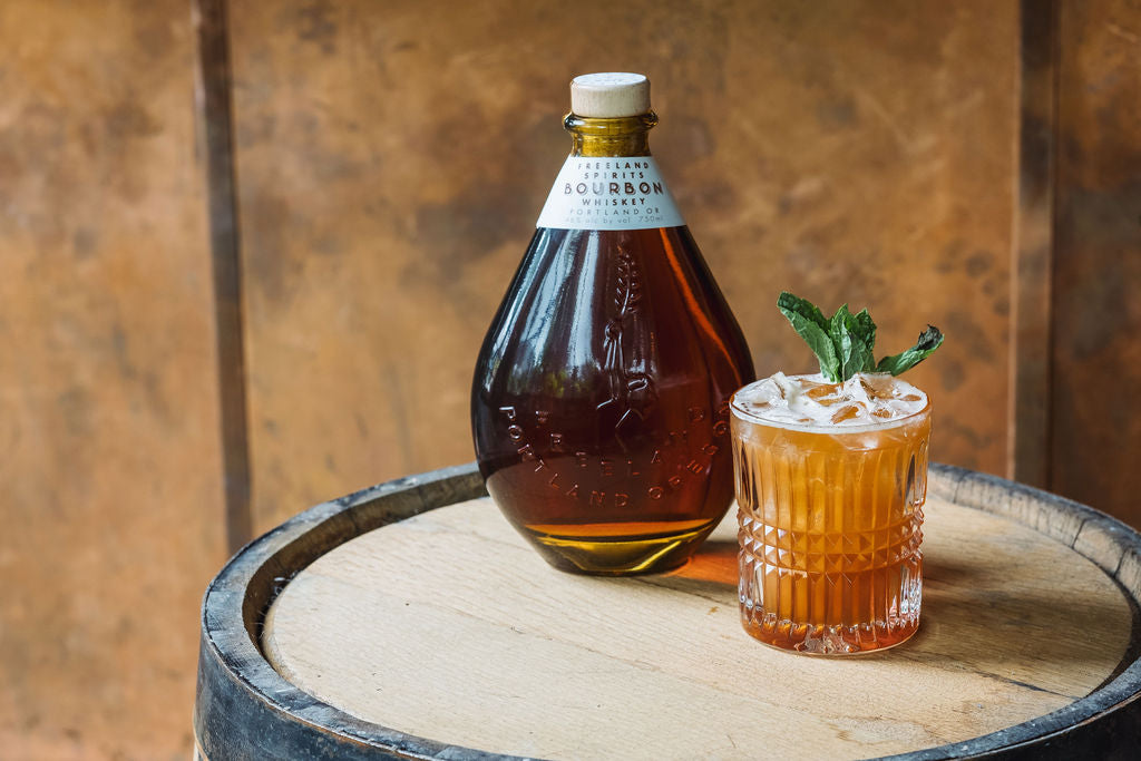 Bottle of Freeland Bourbon on barrel next to Bourbon Mule cocktail garnished with mint