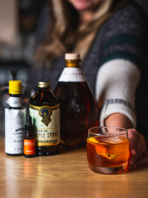 Mixologist serving an Old Fashioned in a branded rocks glass next to the Old Fashioned Kit by Freeland Spirits. Includes Freeland Bourbon, Orange bitters by Portland Bitters Project, Simple Syrup by Portland Syrups, and Angostura aromatic bitters.  