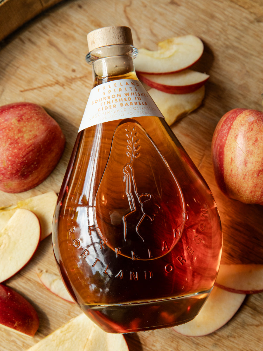 Freeland Spirits Wheated Bourbon Whiskey Finished in Cider Barrels resting on Bauman's Cider barrel surrounded by apples 